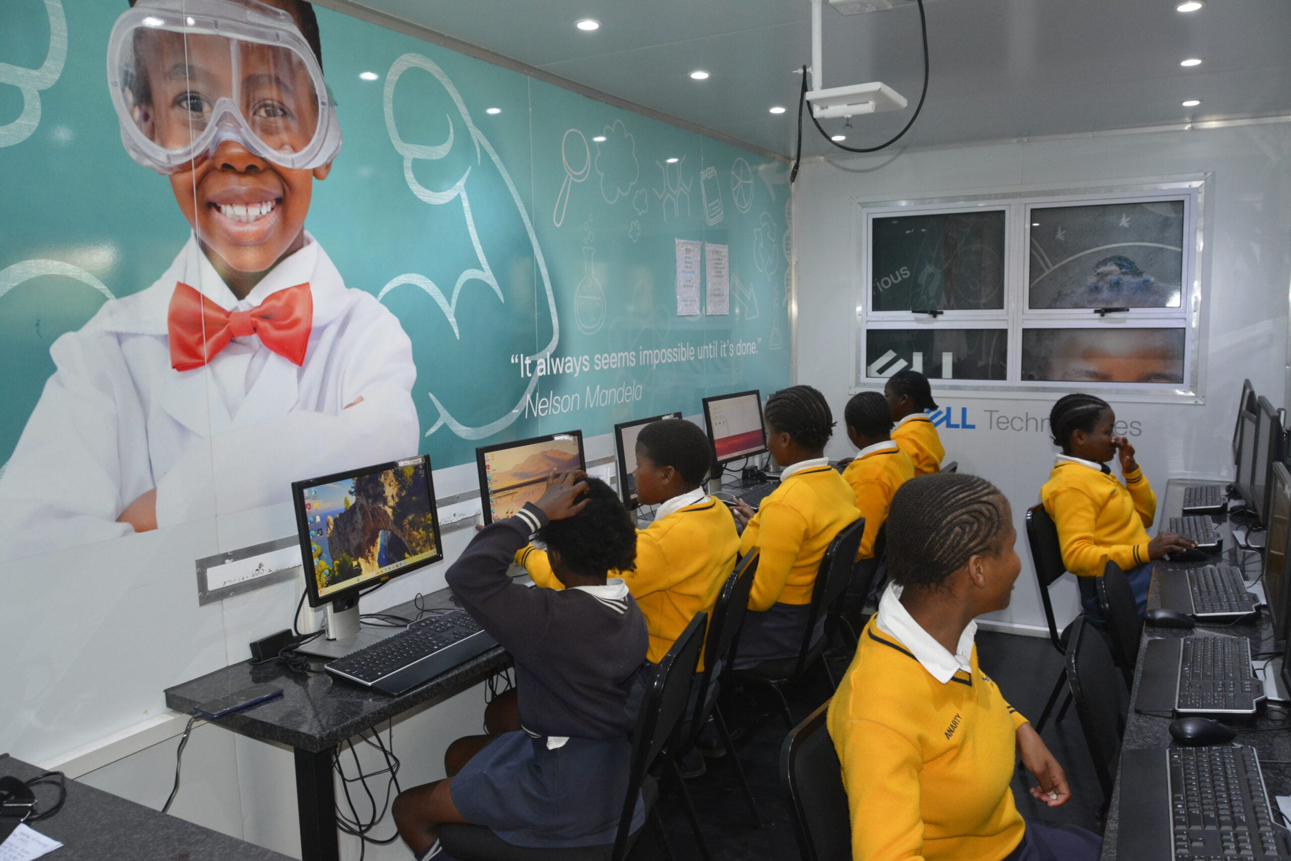 School pupils in yellow uniforms using desktop computers inside a mobile technology classroom.